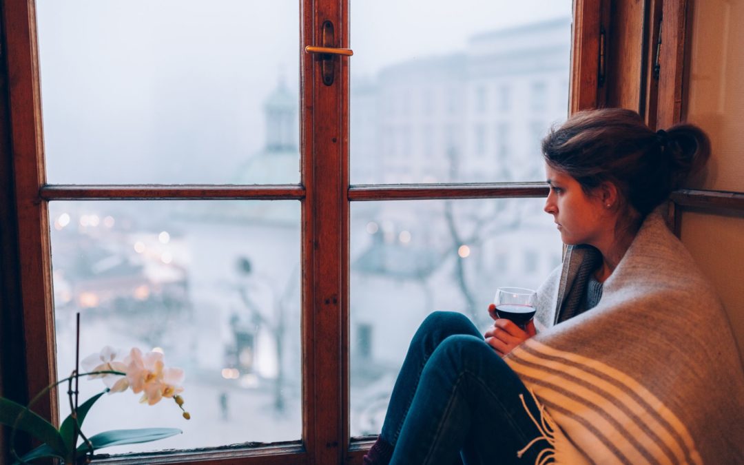 Woman looking out window with blanket wrapped around her.